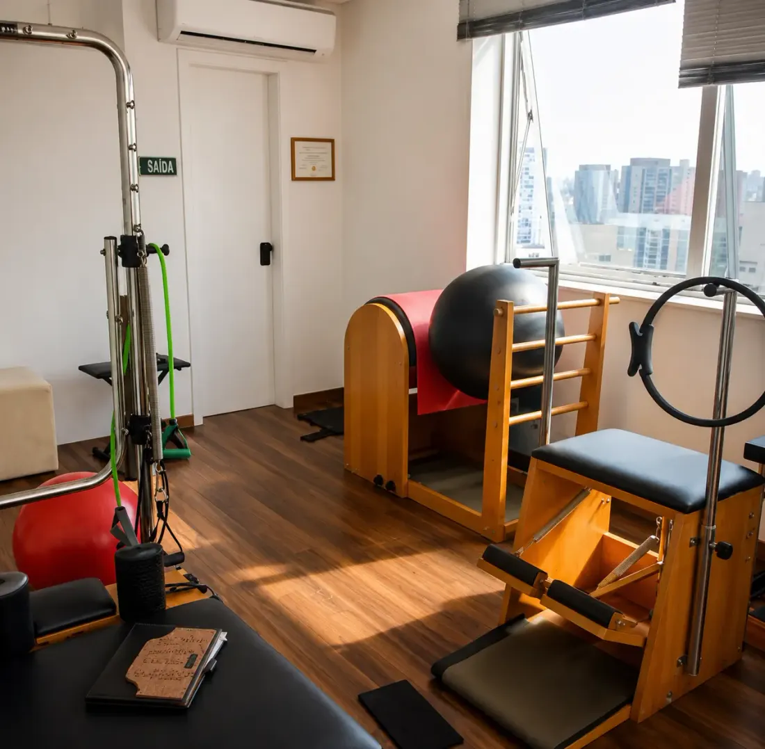 Vista geral do consultório de fisioterapia da Dra. Thelma Soares na Aclimação: equipamentos de Pilates em madeira, luz natural e janelas com vista para São Paulo